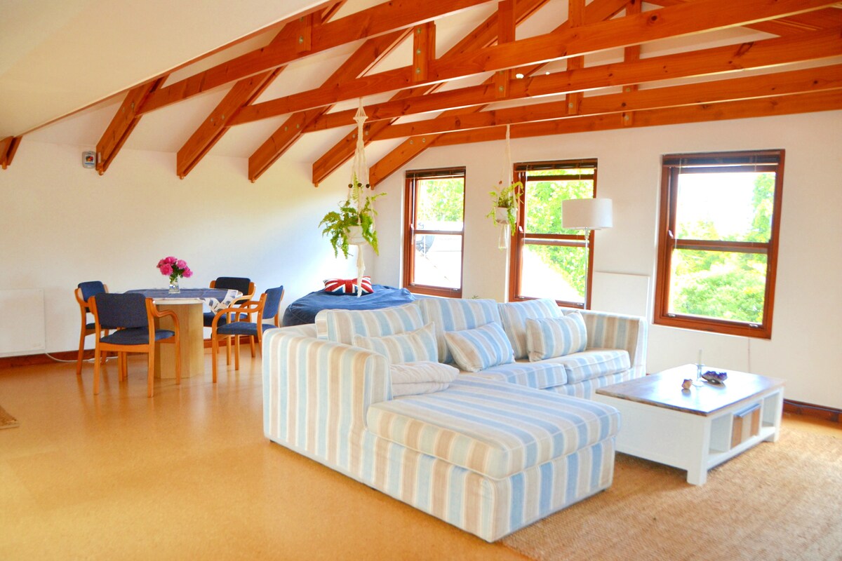 A spacious living area features vaulted wooden ceilings and large windows that allow natural light to fill the room. A comfortable striped sofa sits next to a white coffee table. A dining table with chairs is visible nearby, adorned with a vase of flowers.