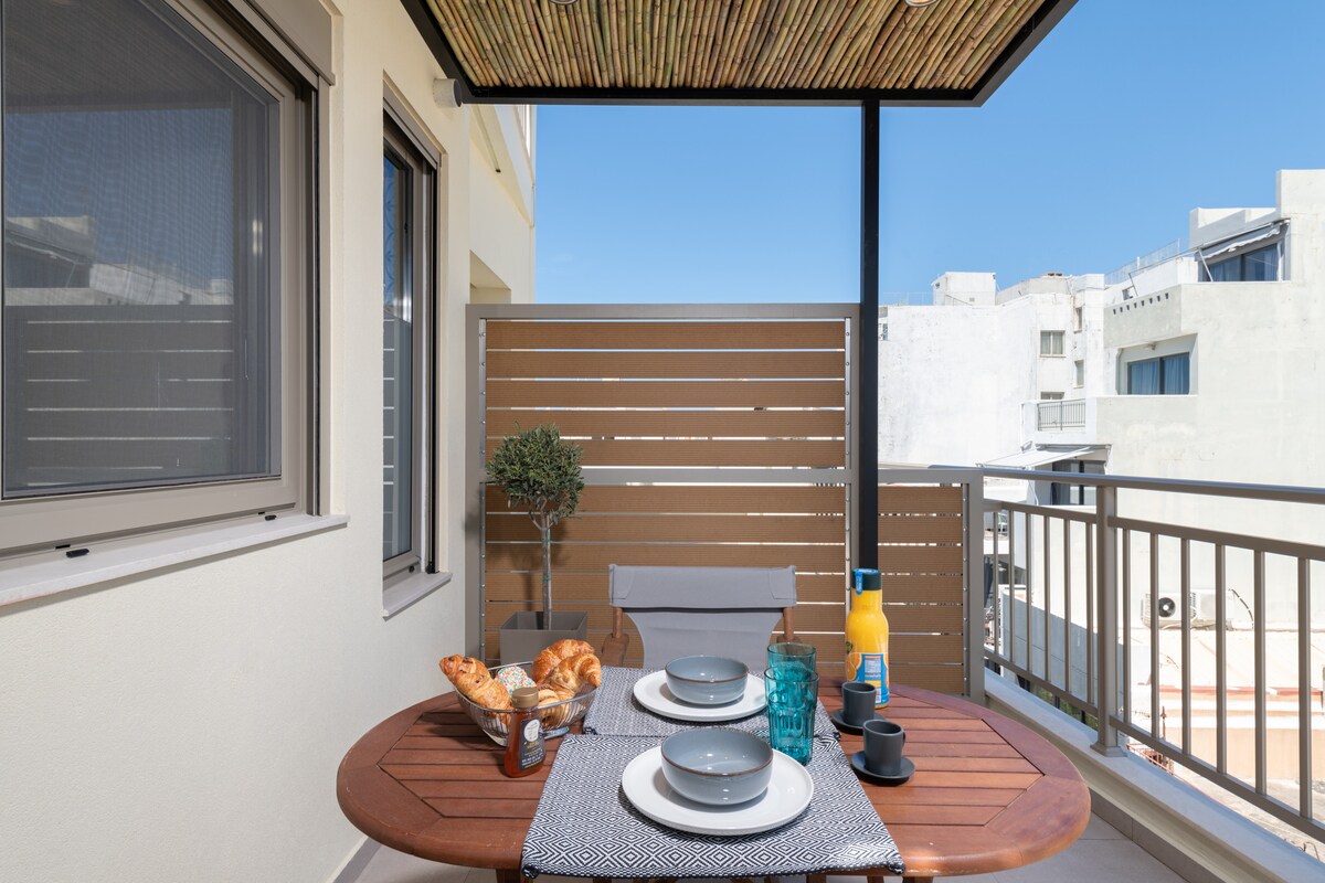 A dining table is set for two on a private balcony, featuring a spread of fresh pastries and drinks. Natural light brightens the compact space, with modern outdoor furnishings and surrounding greenery providing a relaxing environment.
