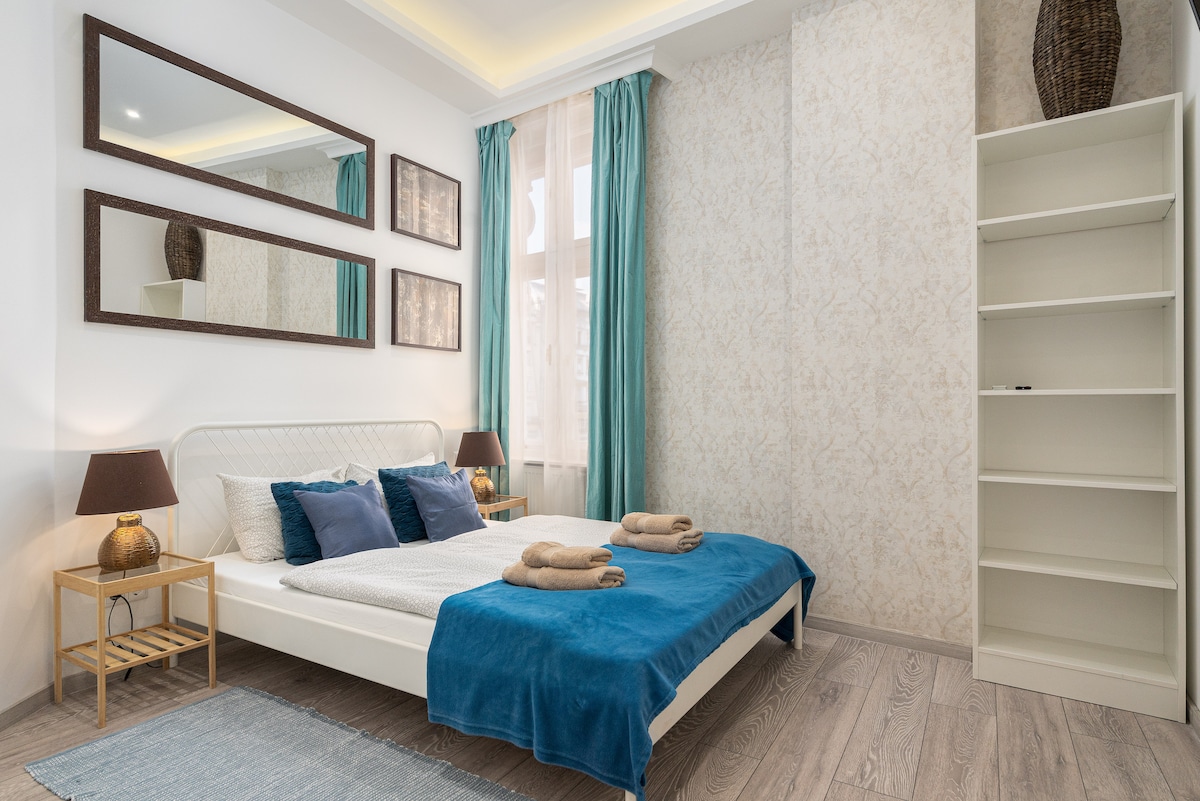 A well-appointed sleeping area features a spacious bed adorned with blue and white linens. Two mirrors reflect the soft lighting, while curtains frame the window. A small shelf and two bedside tables with lamps add functionality to the room, complemented by neatly stacked towels.
