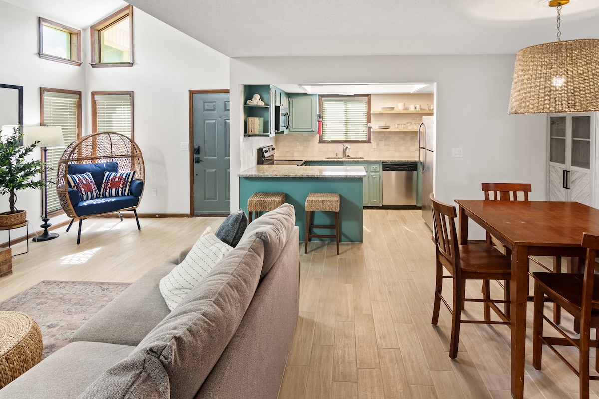 A bright living area features large windows allowing natural light to fill the space. A comfortable sofa faces a wooden dining table, which is surrounded by four chairs. The kitchen, visible in the background, is equipped with modern appliances and a breakfast bar.