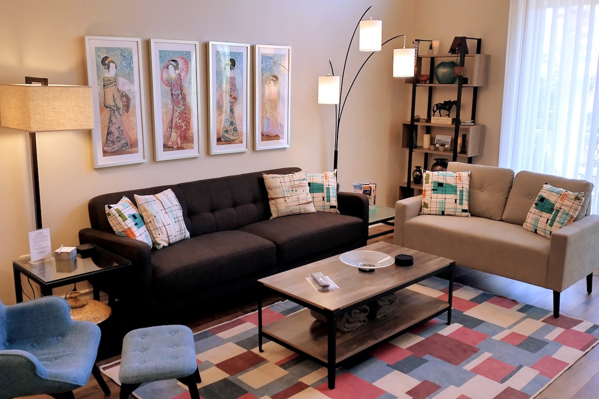 A cozy living area is designed with a contemporary touch, featuring a dark sofa and light-colored loveseat. Colorful throw pillows add interest. A large coffee table sits at the center, and framed art is displayed on the wall. A floor lamp and bookshelf complete the inviting space.