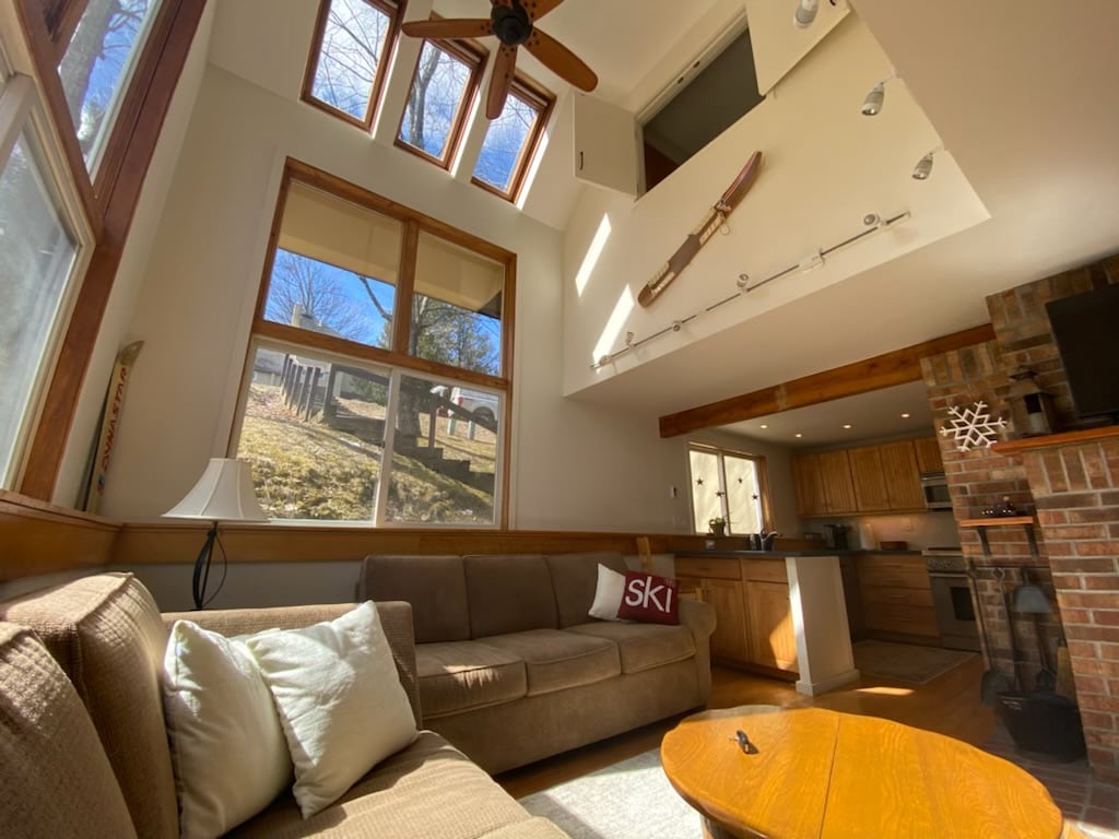 A spacious living room features large windows allowing abundant natural light, showcasing views of the outdoors. A comfortable sofa sits next to a wooden coffee table, while a streamlined kitchen is visible in the background. Decorative ski equipment adds a thematic touch.