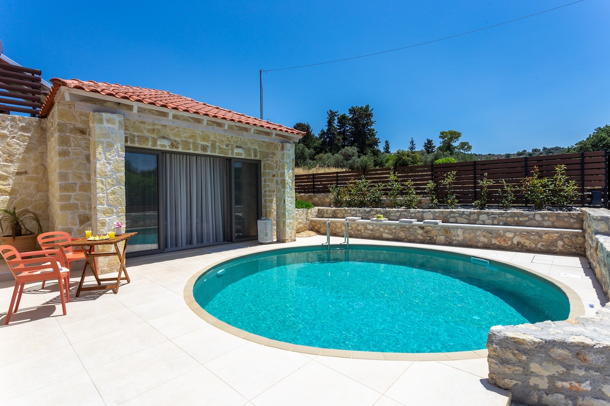 A circular private pool is framed by natural stone walls, surrounded by a tiled patio. Two terracotta chairs and a small table are positioned nearby, providing a place to relax. Large glass doors open into the villa, inviting views of the landscaped garden.