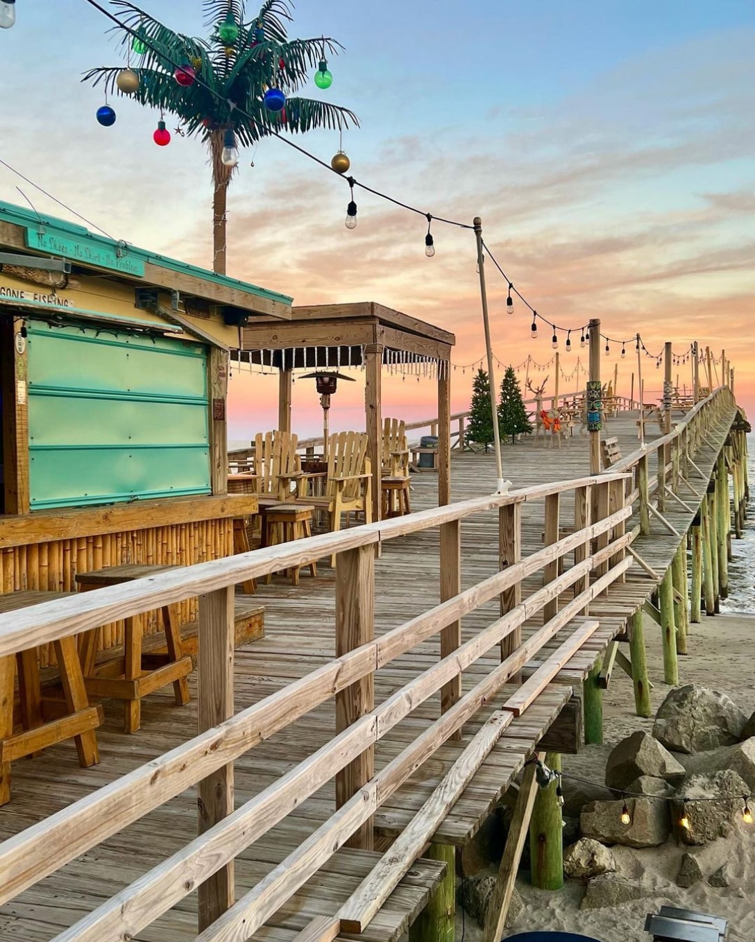 A wooden boardwalk leads towards the water, lined with string lights and a palm tree adorned with colorful decorations. On one side, a casual dining area with wooden benches and tables is visible. The sky displays soft pastel hues as the sun sets.