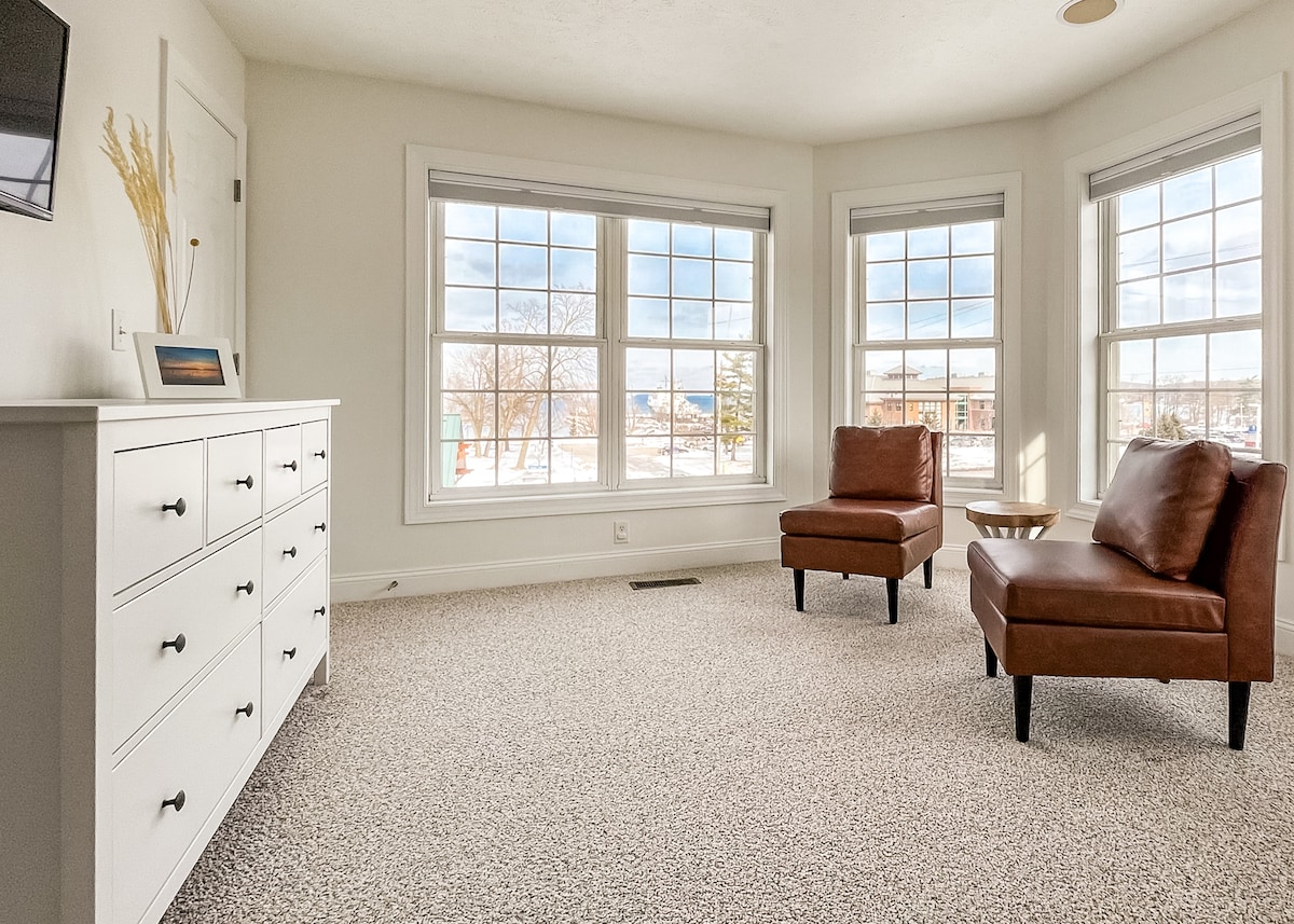 A light-filled living area features two brown leather chairs positioned near large windows, offering views outside. A white dresser is placed against the wall, enhancing the room's neutral palette. Plush carpeting provides a cozy feel to the space.