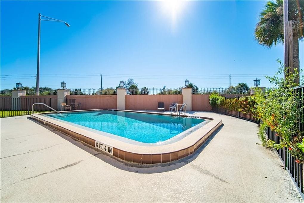 A community pool is seen under bright sunlight, surrounded by a fenced area with mature landscaping. Lounge chairs are positioned near the water, while the pool's gentle curvature adds to its inviting appearance. Clear blue skies create a serene backdrop.