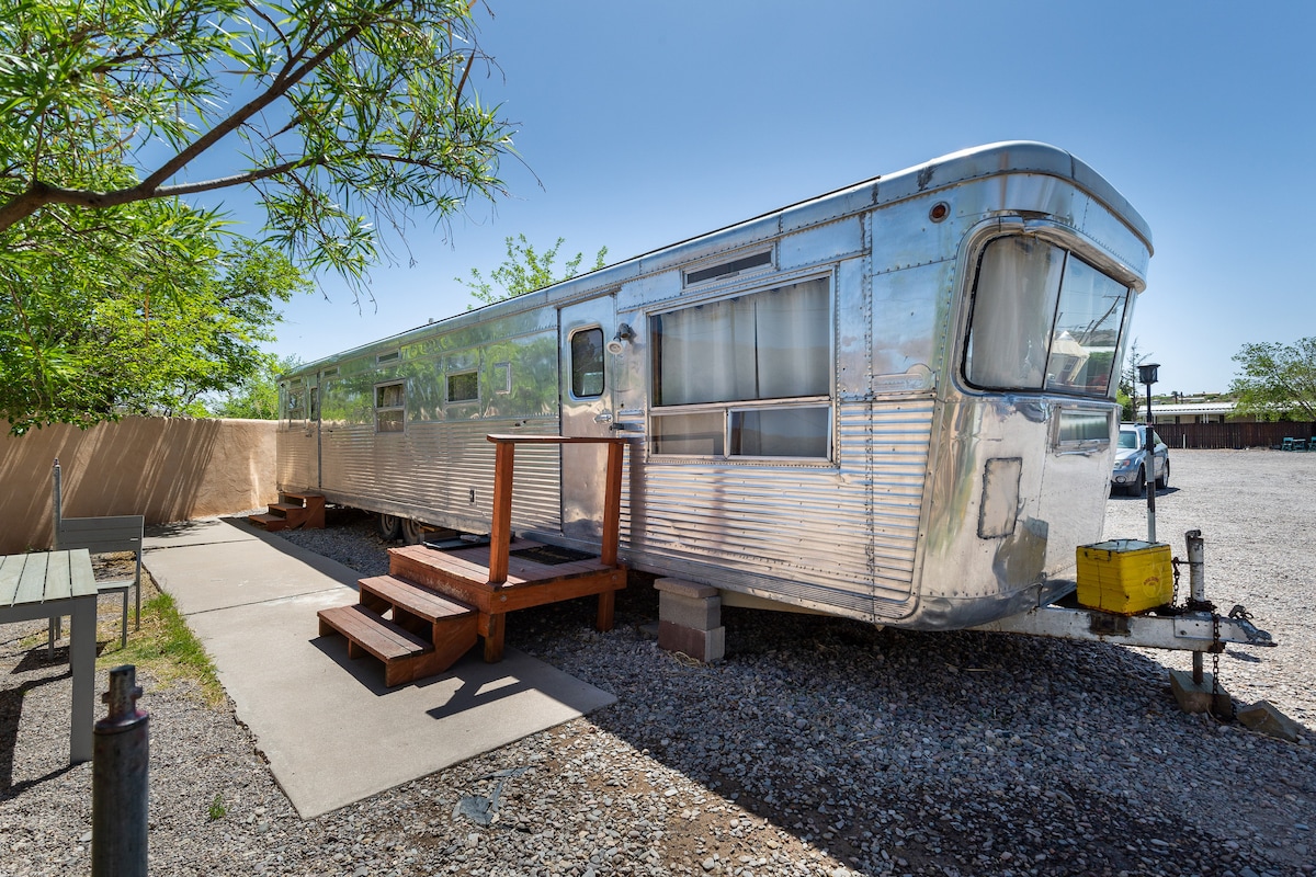 The 1955 Spartan Imperial Mansion glistens under bright sunlight, showcasing its metallic exterior. A wooden deck with steps leads to the entrance, framed by a gravel pathway. Surrounding greenery adds a touch of nature, enhancing the outdoor environment.
