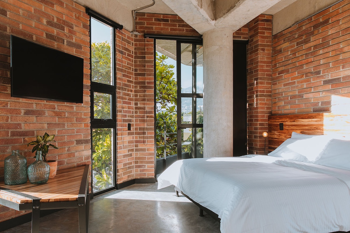 A bright bedroom is highlighted by large windows, offering views of surrounding greenery. A spacious queen bed is positioned against a textured wall of exposed brick. The room features a modern TV mounted on the wall and a wooden table adorned with decorative glass vases.