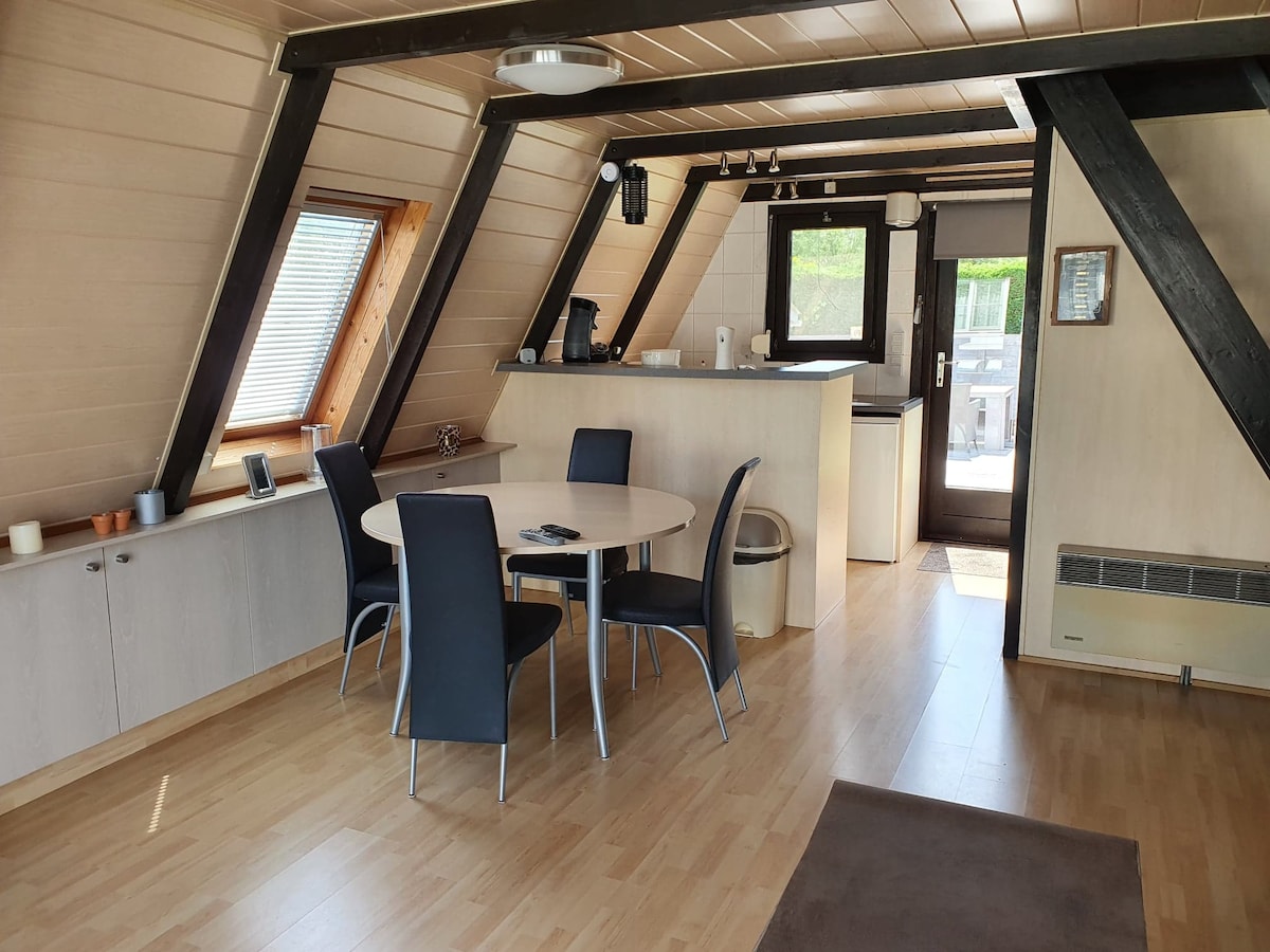 An open dining area features a round table surrounded by four black chairs. A kitchenette equipped with modern appliances is visible in the background, with large windows allowing natural light. The entrance door leads to an outdoor space, enhancing connectivity with the surroundings.