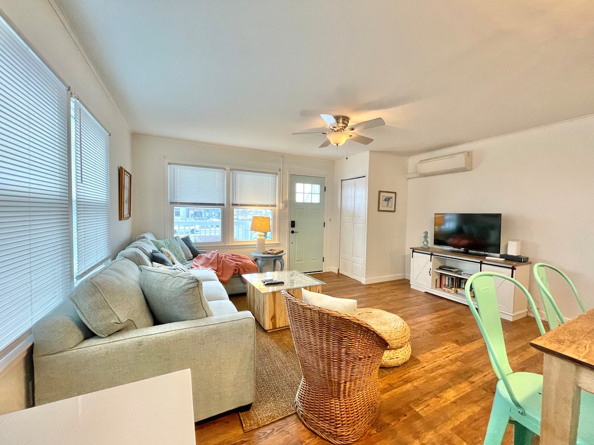 A comfortable living area is presented with a spacious sectional sofa and a cozy armchair. Natural light enters through multiple windows, illuminating the wooden floor. A simple coffee table and a television stand complement the furnishings, creating an inviting space for relaxation.