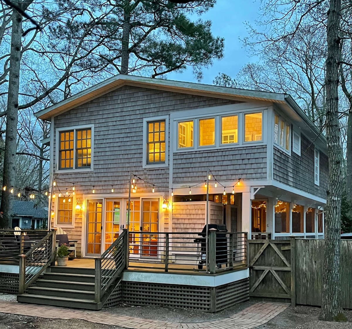 The exterior of a two-story cottage is illuminated by warm light from numerous windows. A spacious deck featuring a grill and surrounded by string lights adds to the inviting setting. The surroundings include a wooden fence and tall trees, contributing to a serene ambiance.