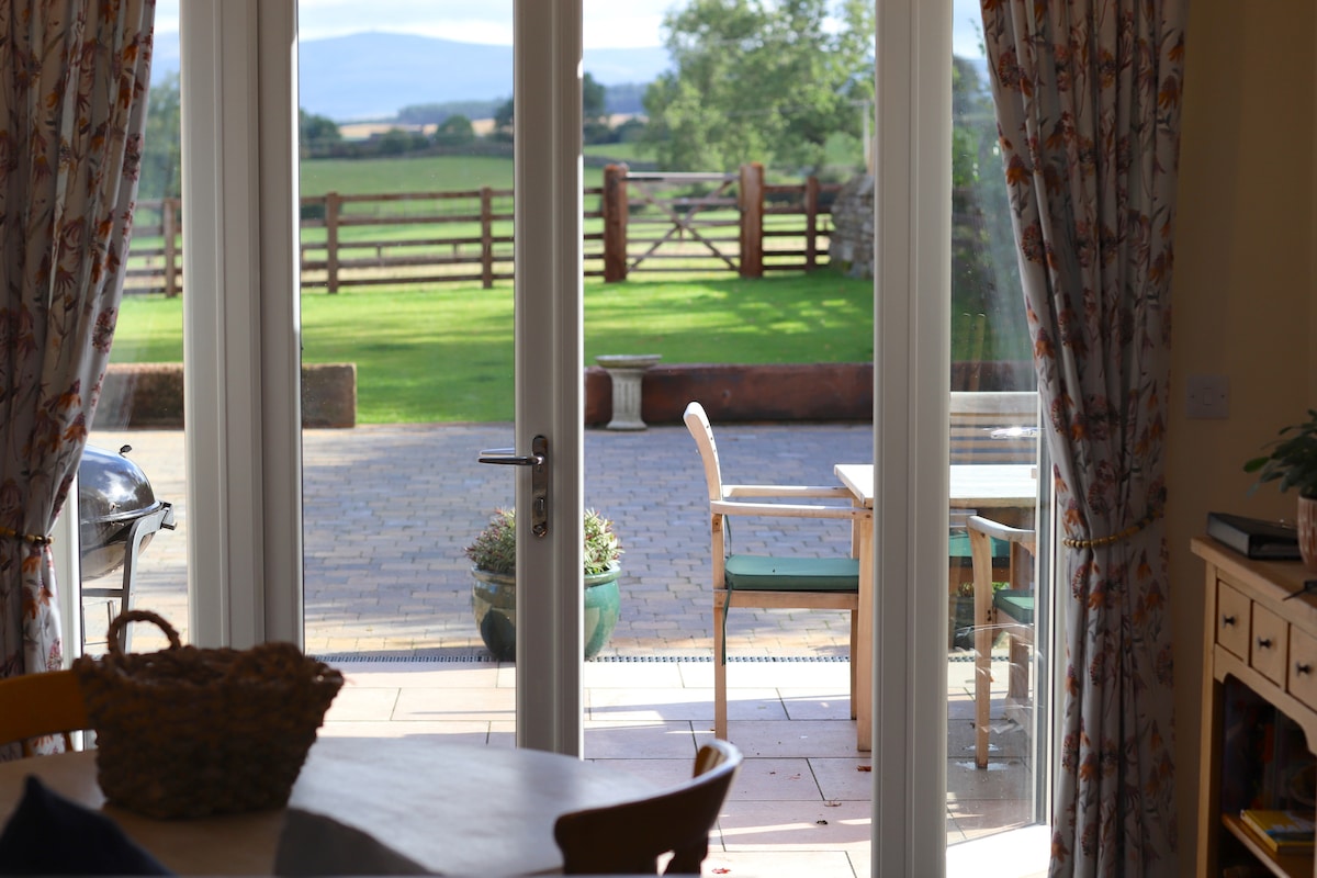French doors open to a spacious terrace with outdoor furniture, overlooking a lawned area and ranch fencing. The scene features a table and chair, with expansive views of the surrounding fields and distant hills.