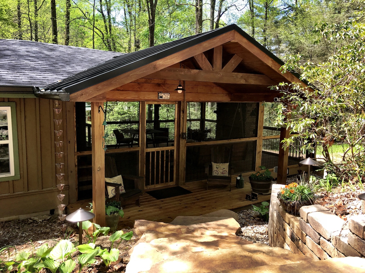 A welcoming cabin entrance features a sturdy wooden porch with a gabled roof. The porch is screened, allowing views of the surrounding woods. Decorative plants are arranged along the pathway, leading to the cabin's inviting entrance.