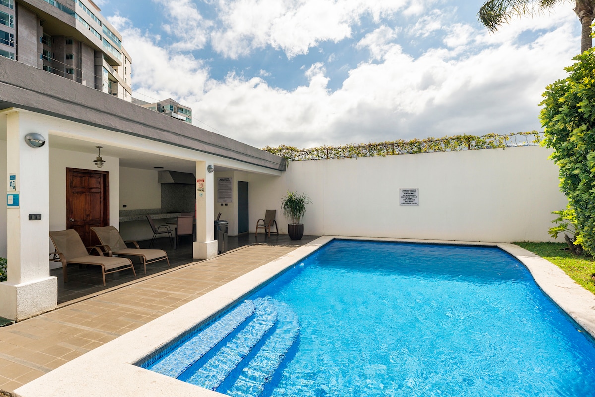 A swimming pool with clear blue water is framed by a patio area featuring lounge chairs. The pool area is complemented by a barbecue space under a covered section, with greenery lining the perimeter, contributing to a relaxed outdoor environment.