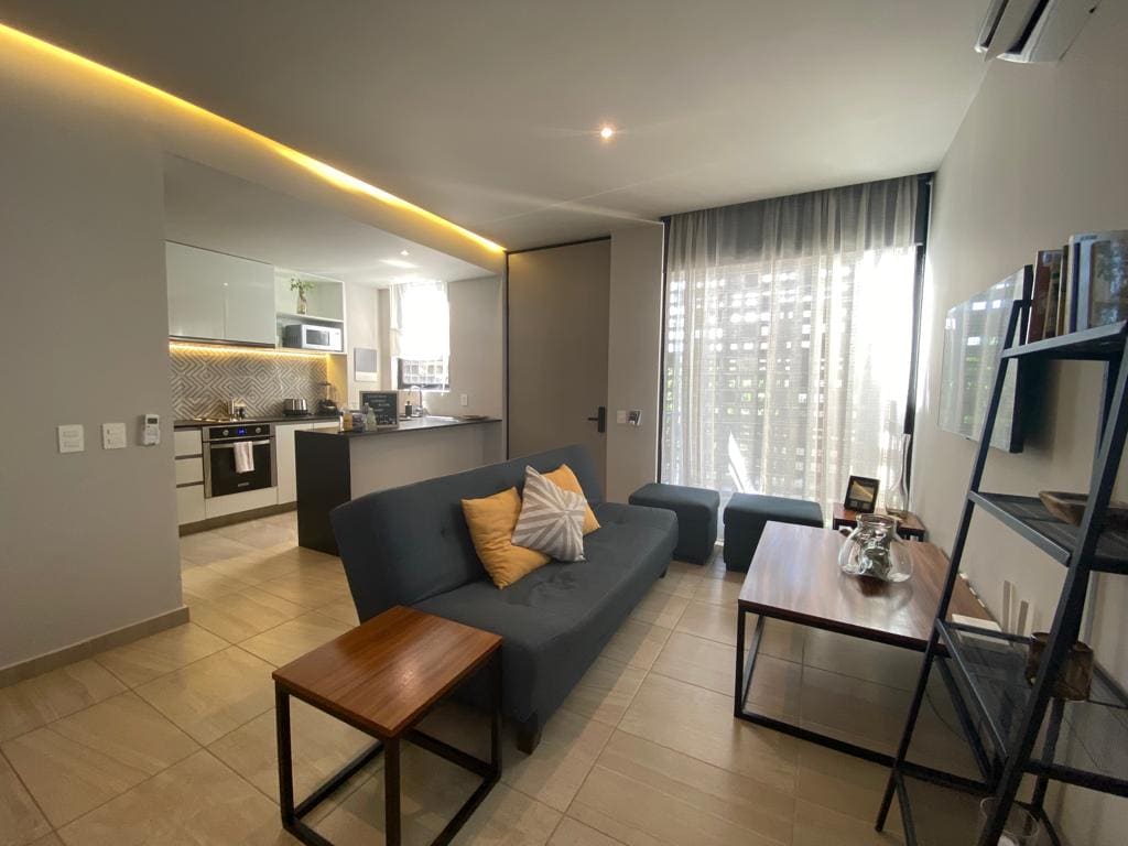 A modern living area is presented with a comfortable dark sofa featuring a decorative yellow pillow. A coffee table and a stool are positioned nearby. The kitchen is visible in the background, showcasing sleek cabinetry and appliances, while natural light filters through the sheer curtains.
