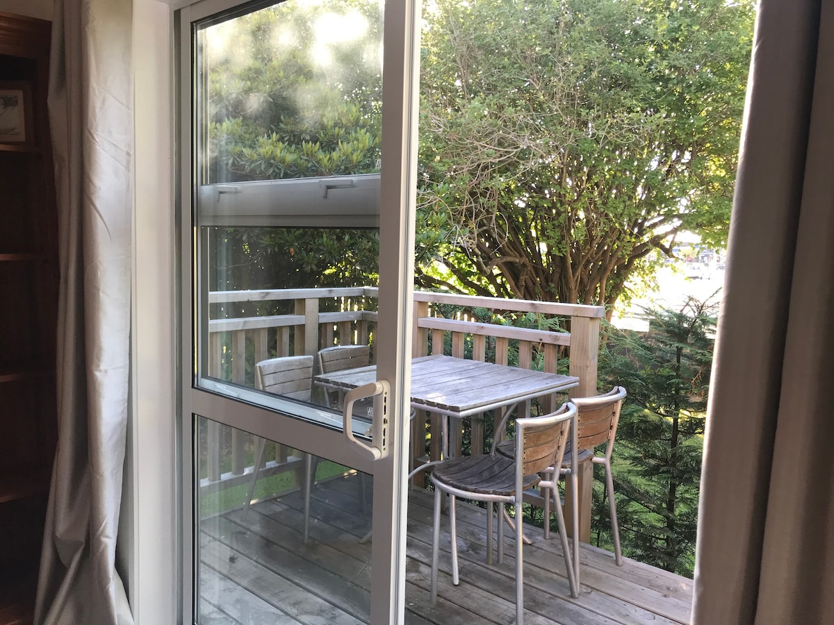 A private deck area is visible through sliding glass doors, featuring a wooden table surrounded by four chairs. Lush greenery frames the view, providing a tranquil setting for outdoor relaxation.