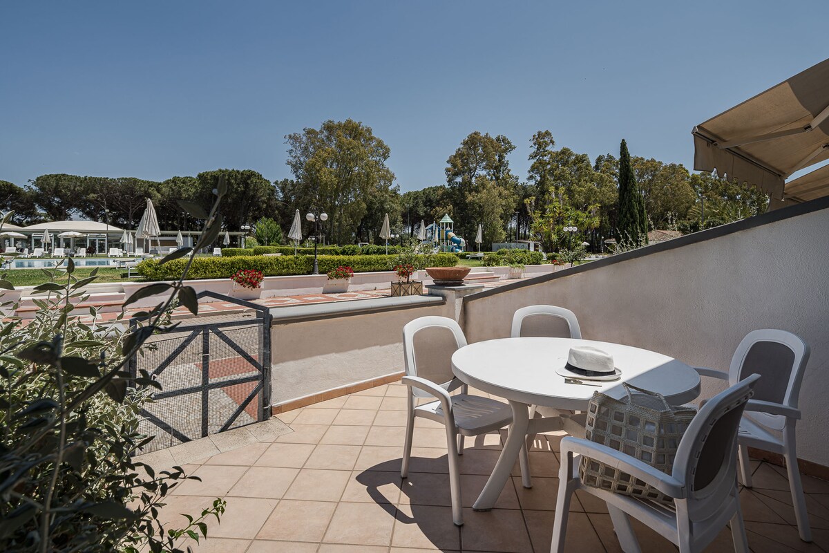 An outdoor terrace features a round dining table surrounded by four white chairs. Potted plants are visible in the background, alongside a pathway leading to a landscaped area with trees and umbrellas, all under a clear blue sky.