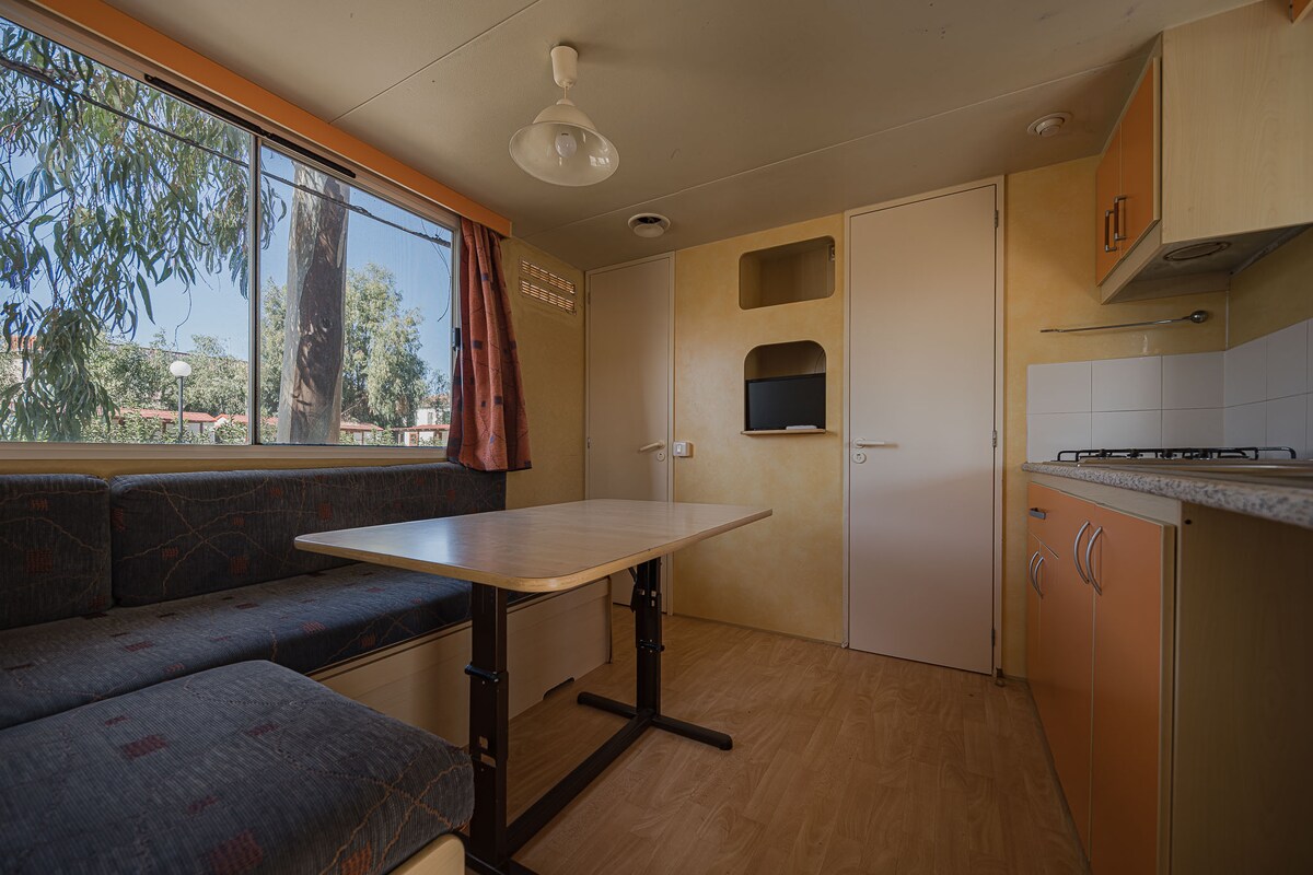 A cozy interior space is illuminated by natural light from a large window, revealing a view of greenery outside. A simple table is positioned near a seating area, with patterned upholstery. A compact kitchenette is visible along one wall, enhancing the functional design.
