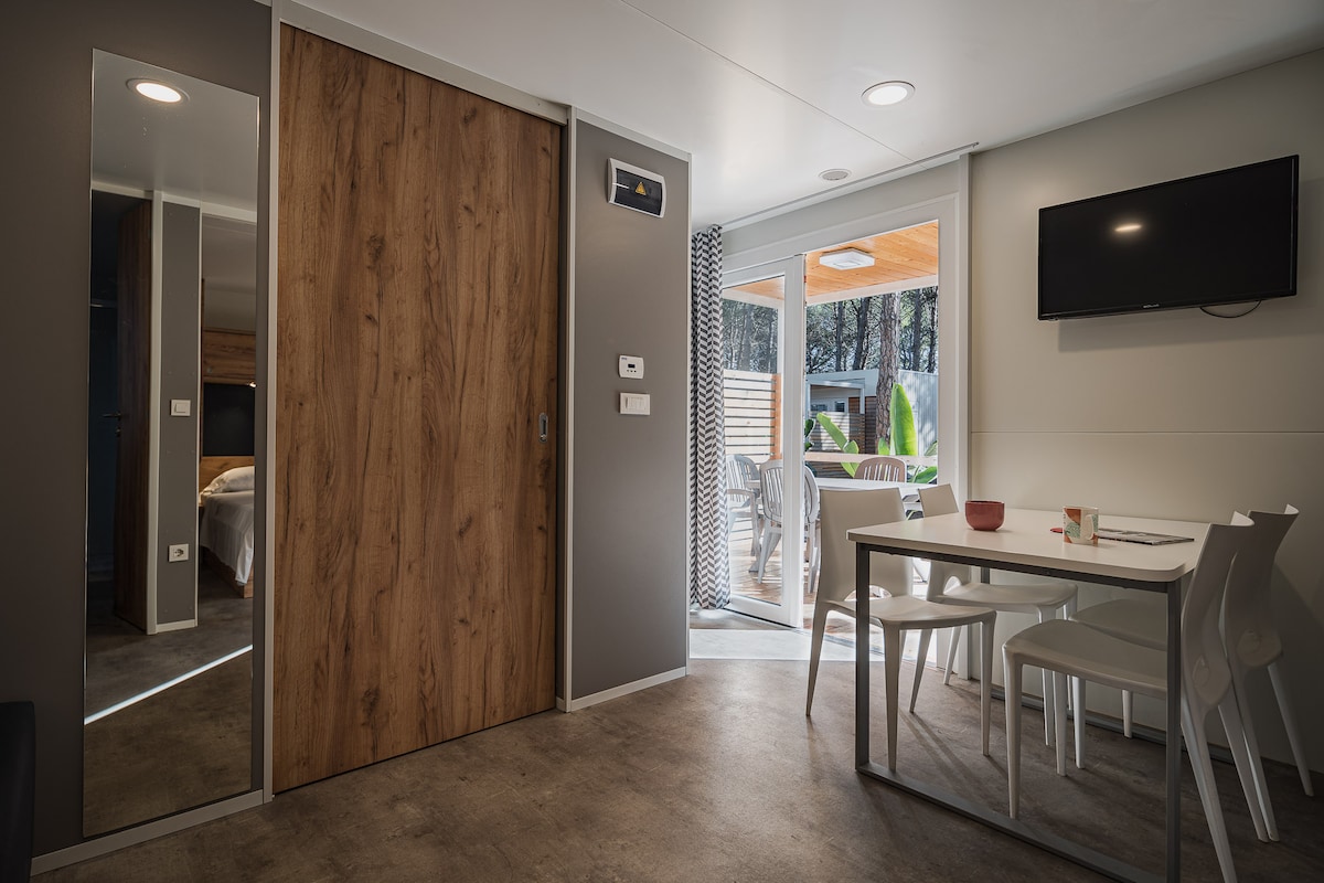 An open plan living area is displayed, featuring a dining table set for four and a wall-mounted television. A sliding wooden door leads to an outdoor terrace, while a full-length mirror adds depth to the space. Natural light filters through large windows.