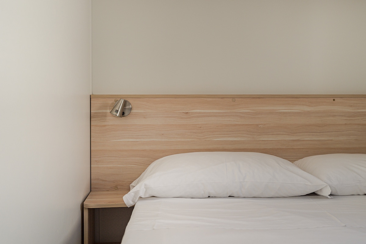 A smooth wooden headboard complements the spacious bed adorned with white pillows. A simple wall-mounted light fixture is positioned above the bed, providing warm illumination against the neutral wall behind.