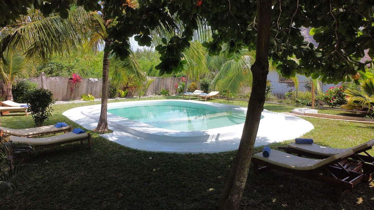 A tranquil swimming pool is surrounded by lush greenery, including palms and flowering plants. Comfortable lounge chairs are positioned nearby, inviting relaxation. The clear water reflects the bright sky, creating a serene atmosphere in the garden.