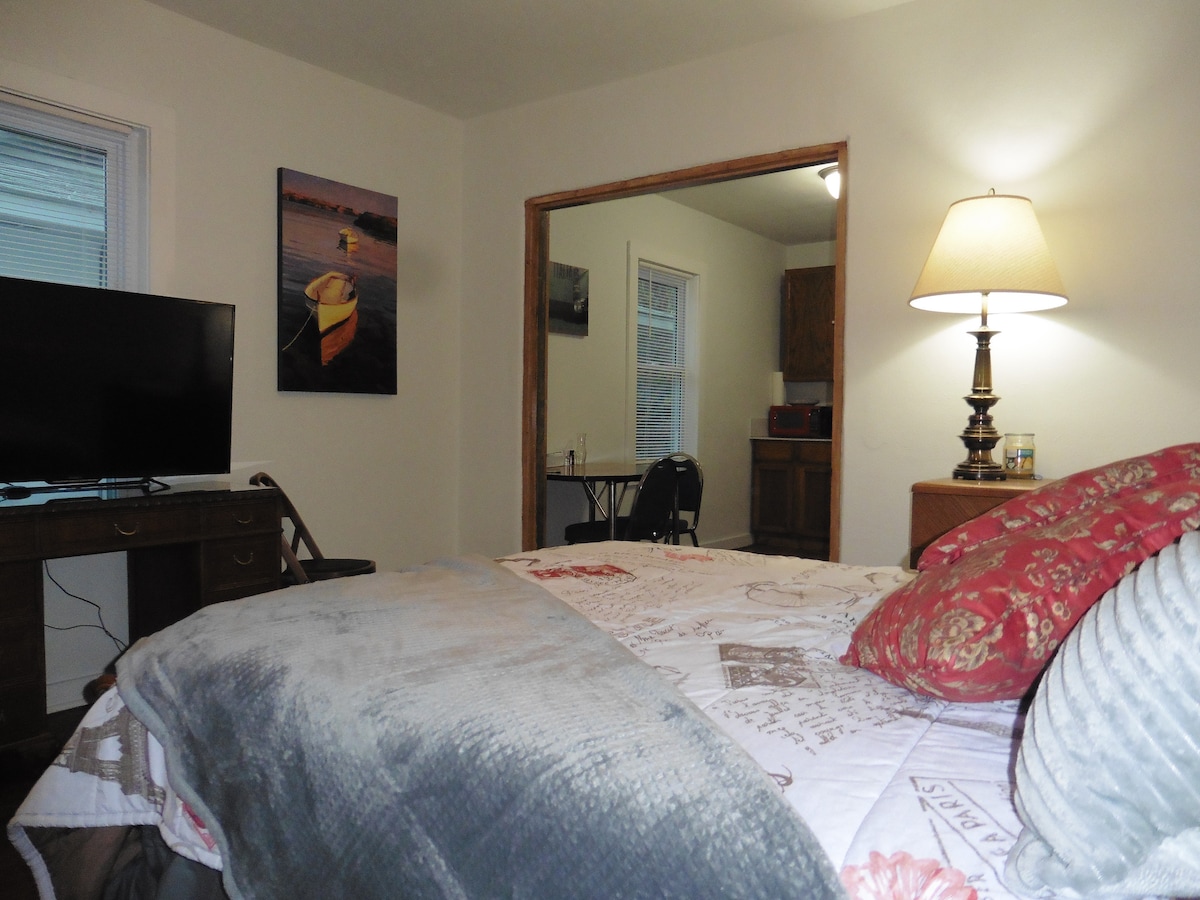 A comfortable resting area is depicted with a large bed layered with decorative pillows and a textured throw. A lamp casts a soft glow beside the bed, while a television and a work desk can be seen in the background, leading into a kitchenette space.