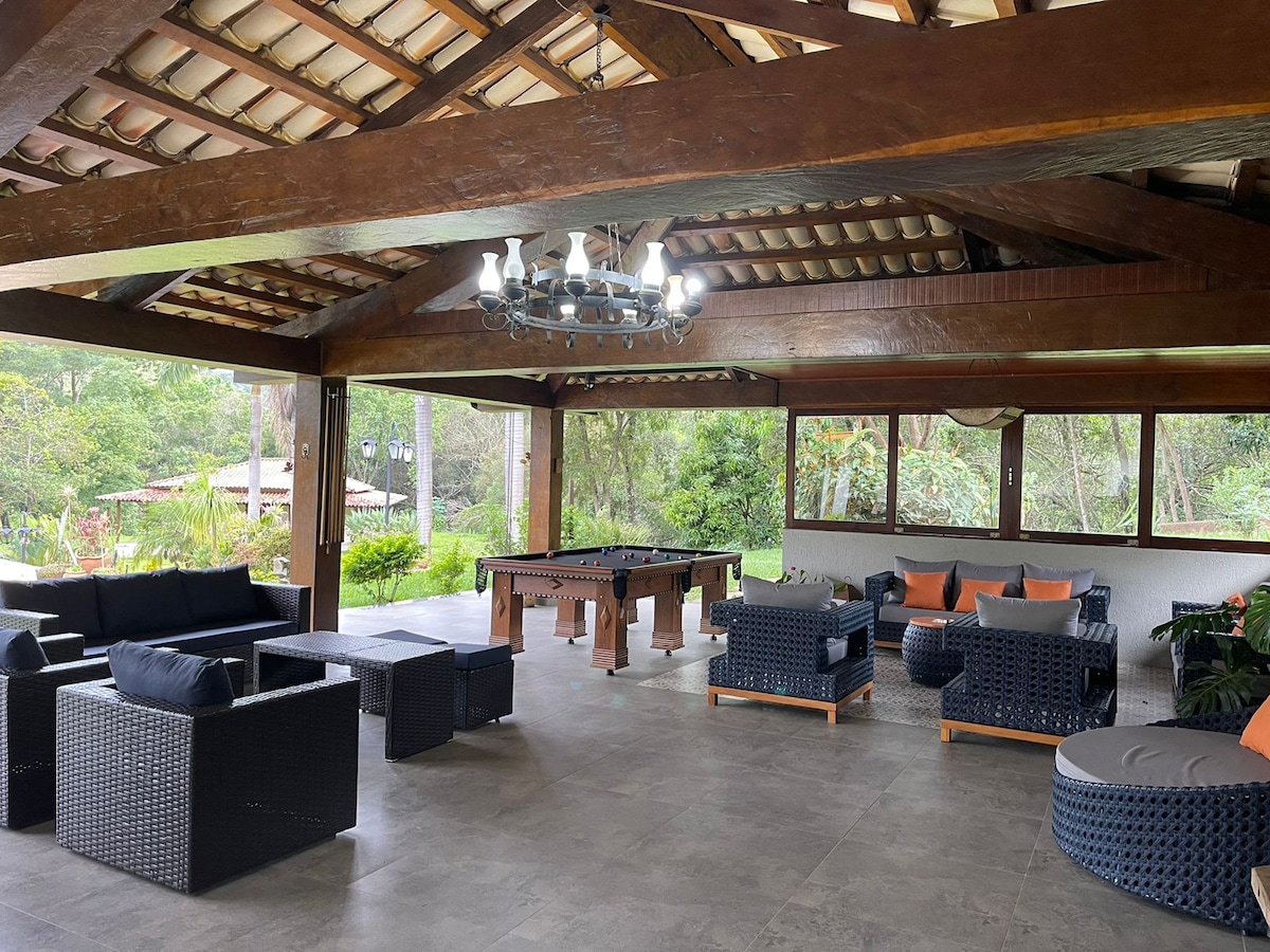 A spacious outdoor lounge area is featured under a wooden beam ceiling. Comfortable seating arrangements made of dark wicker are present, accompanied by bright orange cushions. A billiards table is visible in the center, surrounded by lush greenery outside.