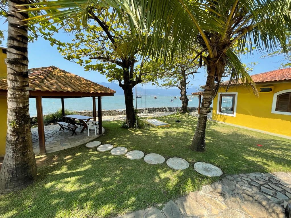 An outdoor area is showcased with a vibrant yellow house in the background. A shaded gazebo is visible, featuring a wooden table and benches, surrounded by lush greenery. The ocean can be seen in the distance, with small boats anchored near the shore.