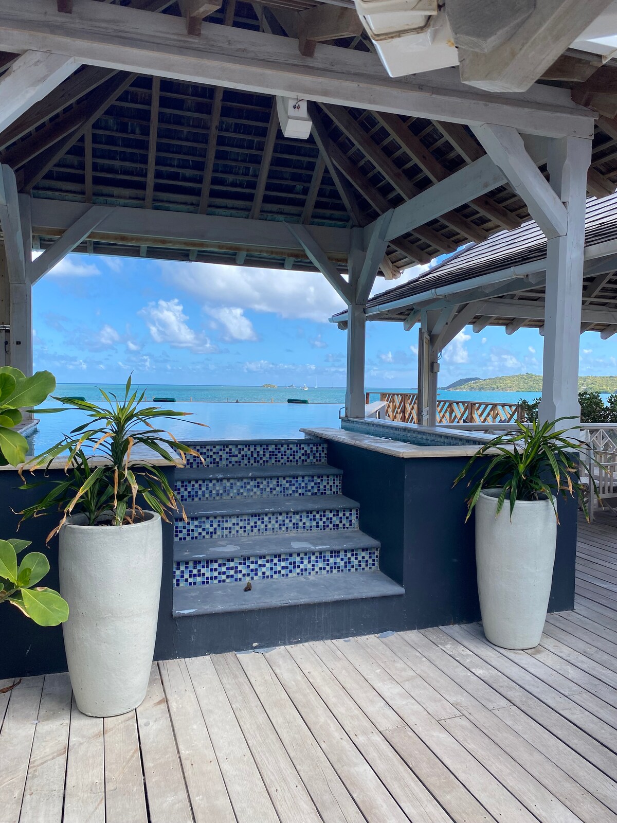 A shaded gazebo is presented with a view overlooking the water. Steps lead down from the gazebo, flanked by tall, white planters holding lush green plants. A wooden deck provides a spacious area for relaxation and enjoying the coastal scenery.