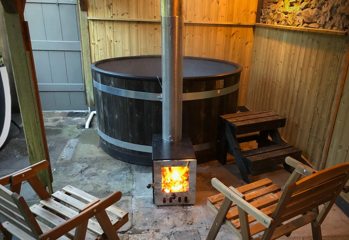 A wooden hot tub is centered in a rustic space, surrounded by wooden walls. A metal chimney rises from a heater, warming the area. Two wooden chairs with slatted seats are positioned beside the hot tub, inviting relaxation in a tranquil setting.