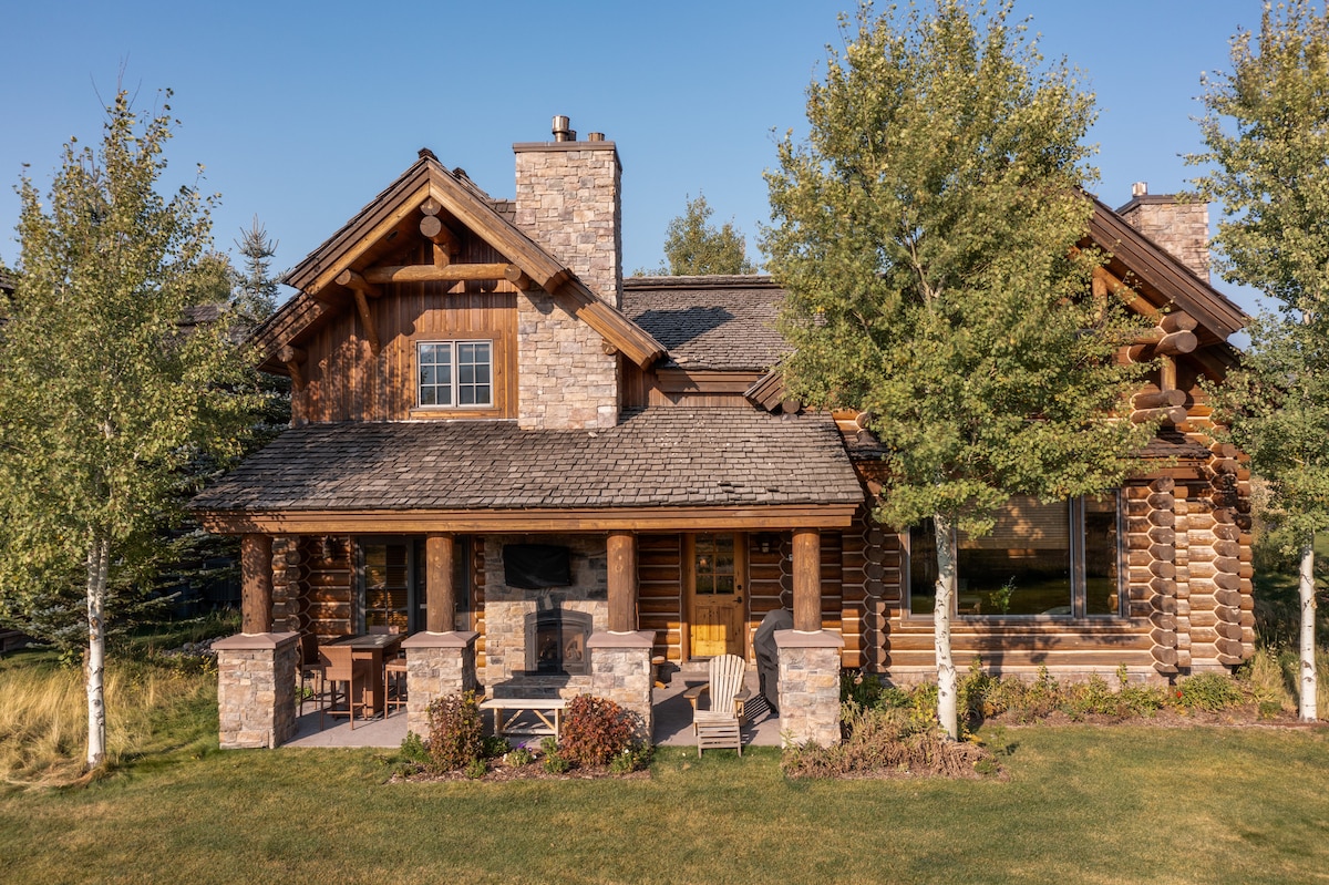 The exterior of a custom log home is shown, featuring a combination of stone and wood construction. A covered porch with wooden chairs overlooks a landscaped yard with shrubs and trees. The structure is framed by clear blue skies, highlighting its rustic charm.
