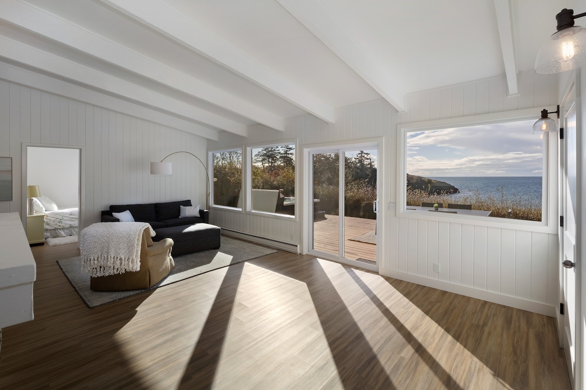A sunlit living area features large windows that frame views of the waterfront. A dark sofa is complemented by a blanket, while light wood flooring extends throughout. The open layout connects to a deck, enhancing the spacious layout of the room.