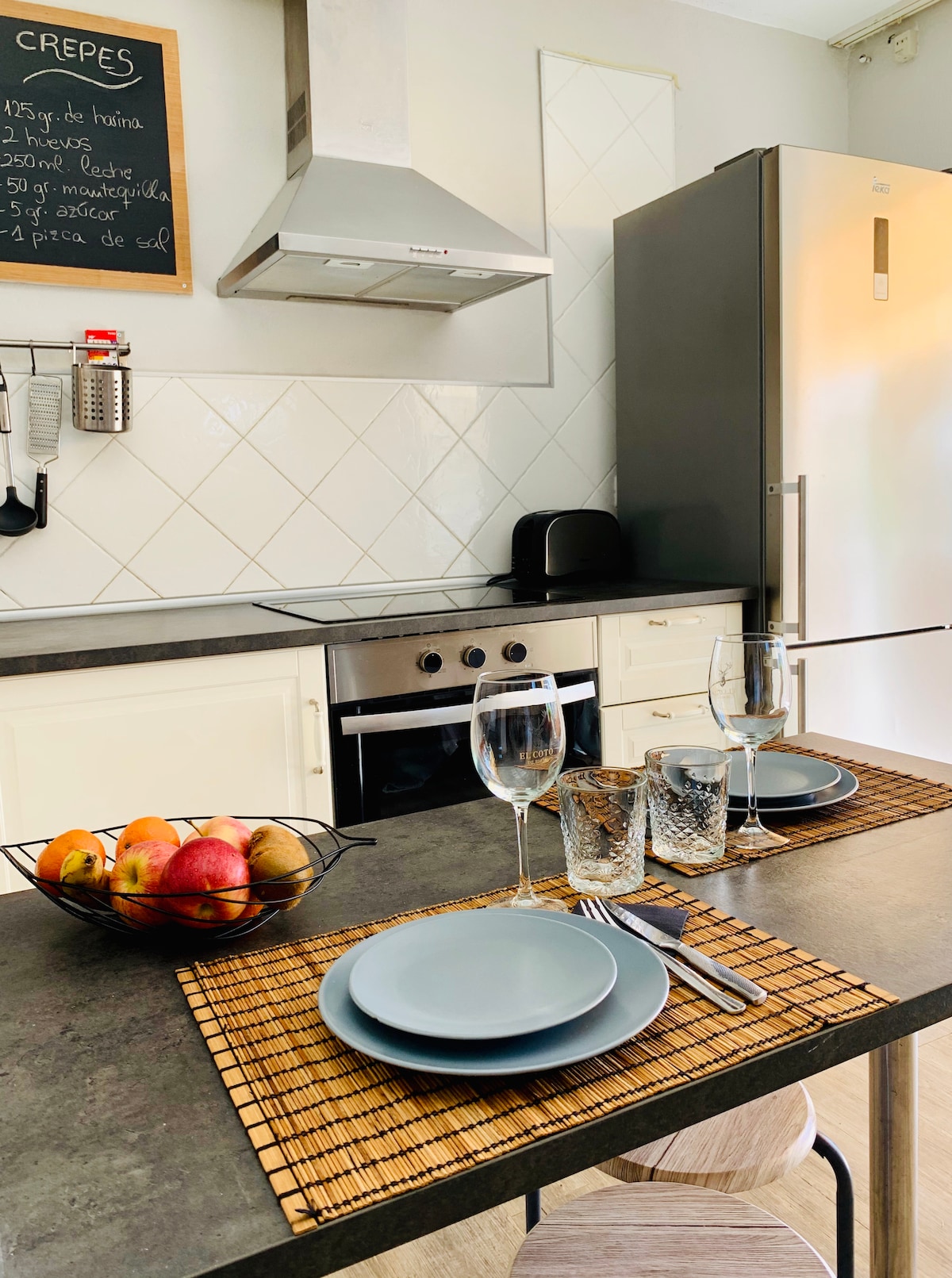 A modern kitchen space is equipped with stainless steel appliances, including an oven and refrigerator. A dining area features a dark countertop with plates, glasses, and a bowl of fruit. A bamboo mat adds texture beneath the settings.