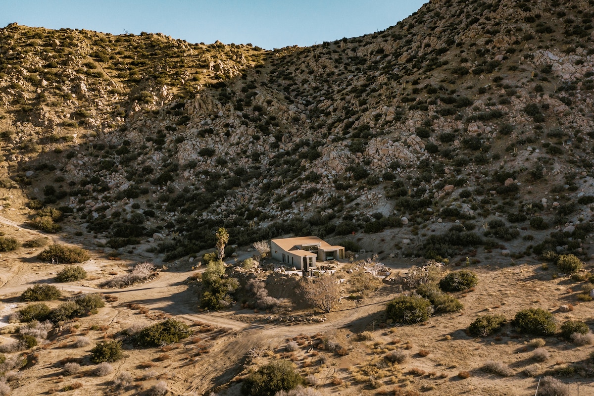 A modern structure nestled in a natural desert landscape is captured from an elevated perspective. Surrounding rocky hills and sparse vegetation create a serene backdrop, emphasizing the remote and tranquil setting of the property.