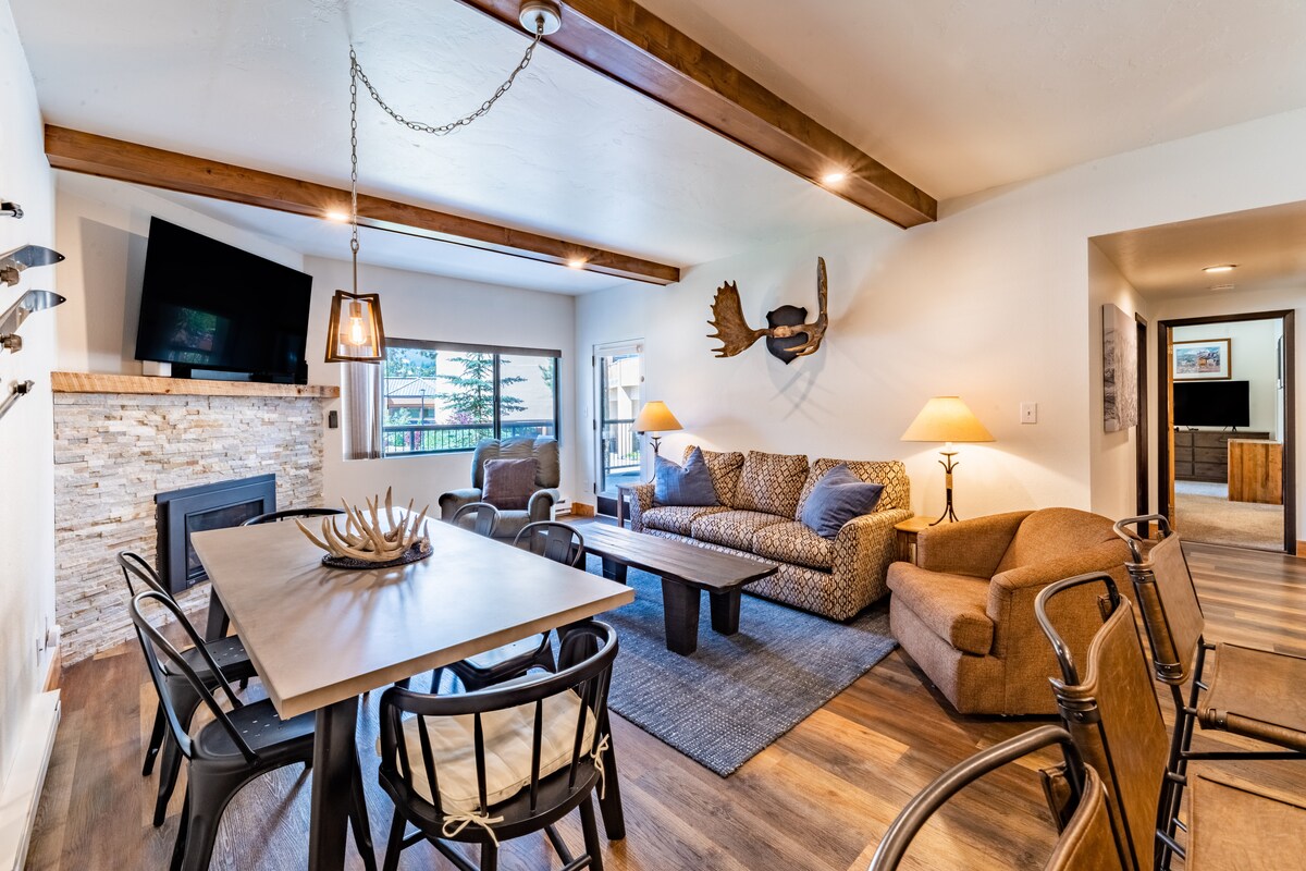 An open living area features a stone fireplace surrounded by a comfortable sofa, armchairs, and a dining table with six black chairs. Natural light fills the space through large windows, while wooden beams add a touch of character to the ceiling.