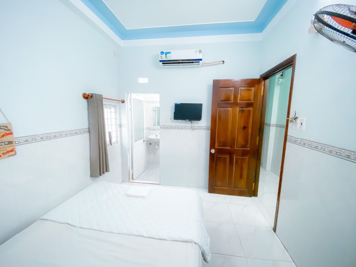 A neatly arranged room is presented, featuring a spacious bed with crisp white linens. A wall-mounted TV is visible, alongside an air conditioning unit and a fan. Natural light filters in through a nearby window, while a light blue ceiling adds a subtle touch of color.