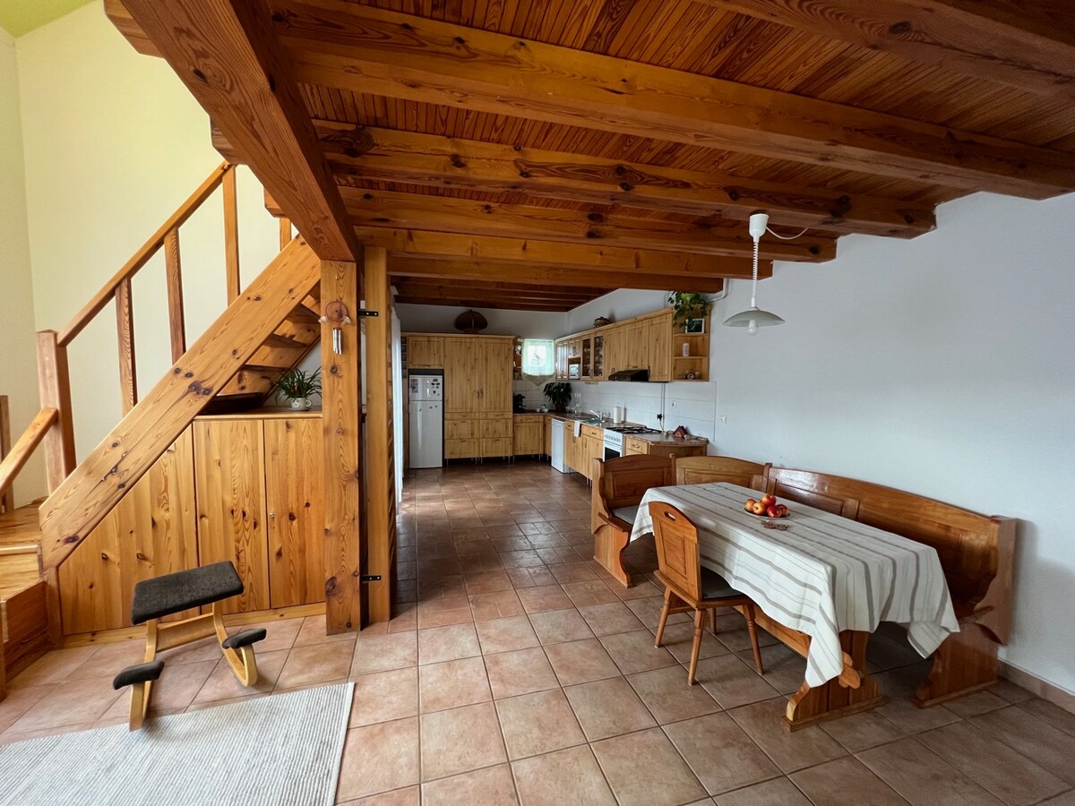 A spacious dining area features a wooden table surrounded by benches and a chair, with a light tablecloth draped over it. The warm wooden beams of the ceiling are complemented by tiled flooring, while a staircase leads to an upper level. A simple kitchen is visible in the background.