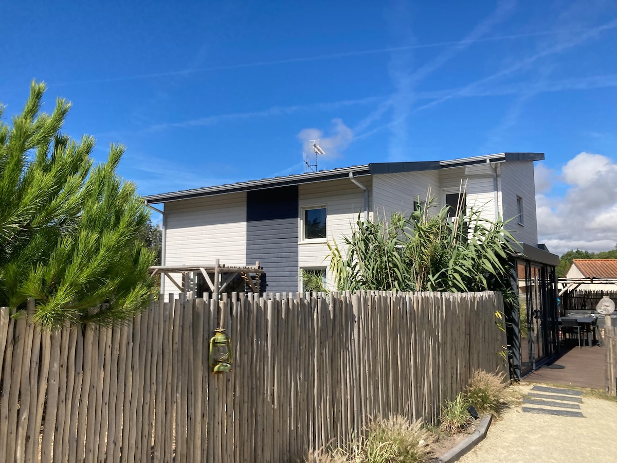 The exterior of the villa is showcased, highlighting a modern two-story structure surrounded by greenery. A wooden fence provides privacy, while natural pathways lead to the entrance. The sky above is clear with soft clouds, contributing to a serene atmosphere.