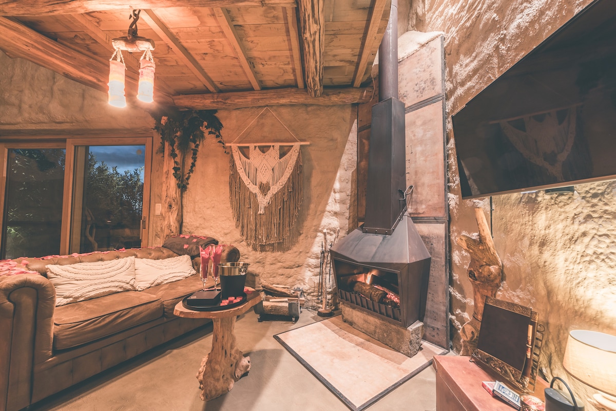 The living area features a cozy brown sofa and a rustic coffee table. A modern fireplace is integrated into the natural stone wall, enhancing the warmth of the room. Large windows allow for ample natural light, with decorative elements adding a tranquil touch.