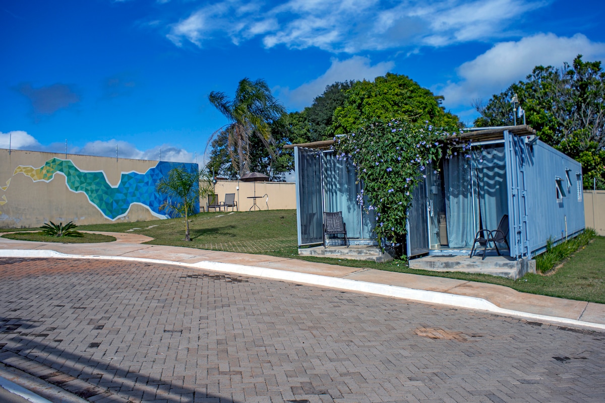 A landscaped outdoor area is shown, featuring two insulated containers. They are positioned on a paved road, surrounded by greenery and a mural wall in the background. Two chairs are placed in front of the containers, inviting relaxation.