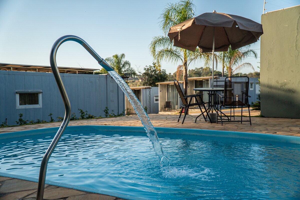 A clear blue pool with a sleek metal ladder gently spills water, creating a calming sound. In the background, palm trees sway, complementing a shaded seating area with a table and chairs under an umbrella.