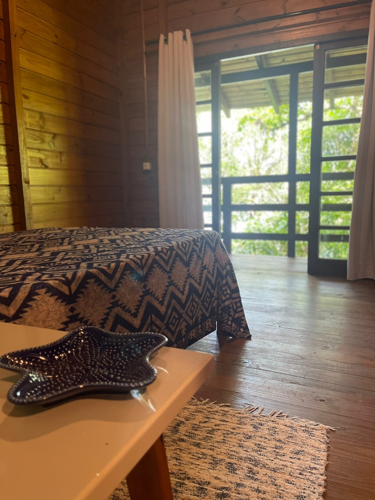 A wooden interior space features a patterned bedspread on a spacious bed. Large glass doors provide access to a balcony surrounded by greenery, allowing natural light to illuminate the room. A decorative star-shaped dish is placed on a nearby table.