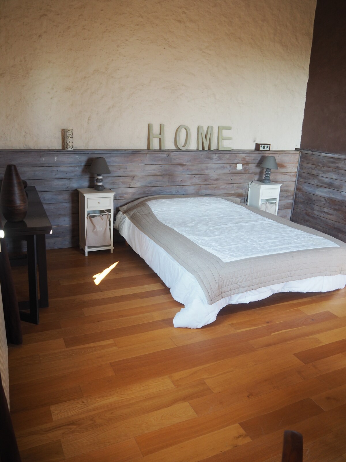 A spacious sleeping area features natural wood flooring and a calming color palette. A light quilt covers the bed, accompanied by two bedside tables with lamps. Decorative letters spell 'HOME' on the wall above, contributing to a comfortable atmosphere.