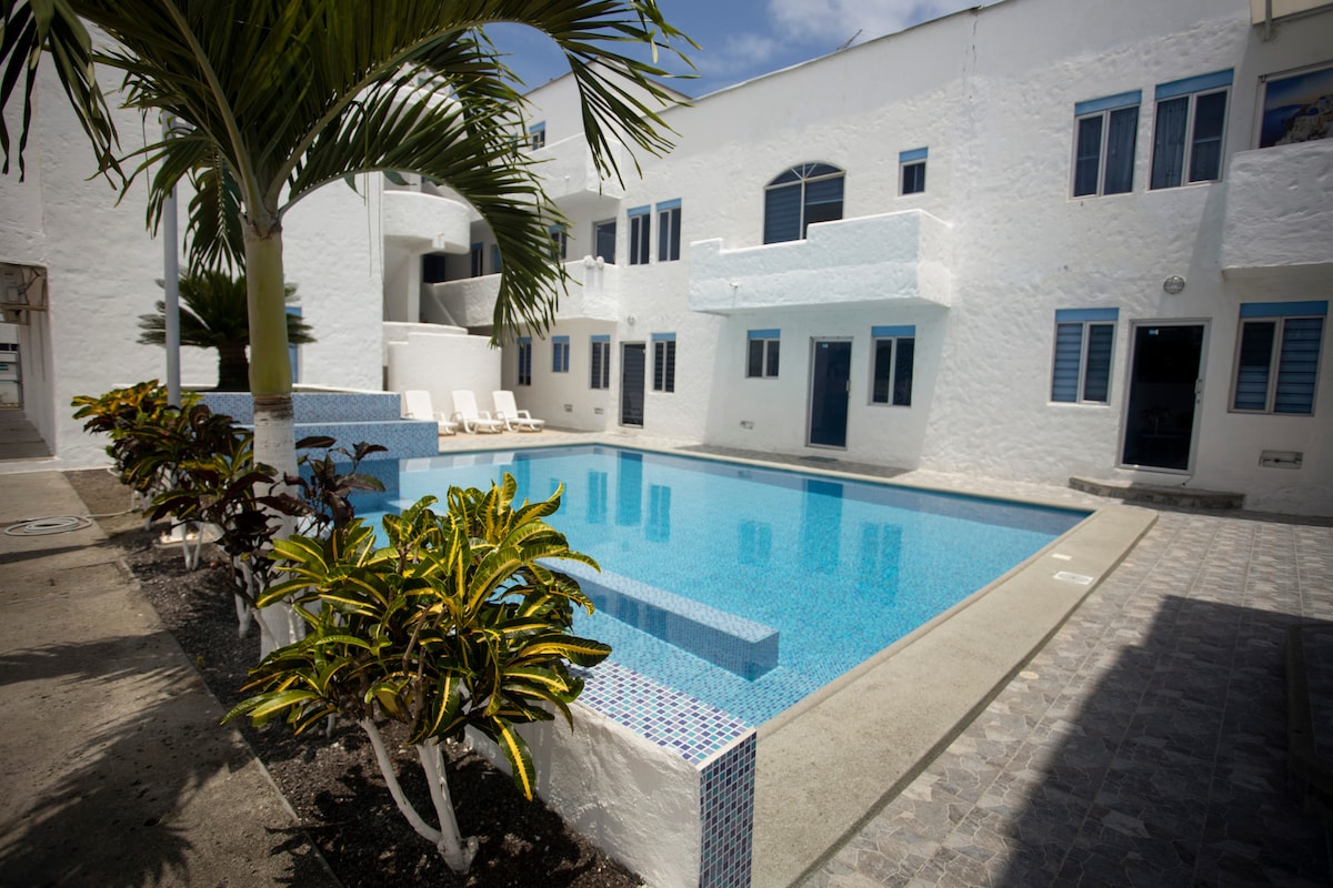 A clean swimming pool is set in the courtyard of a bright white building. The surrounding area features tropical plants and lounge chairs, inviting relaxation. Large windows and doors of the building can be seen, enhancing the openness of the space.