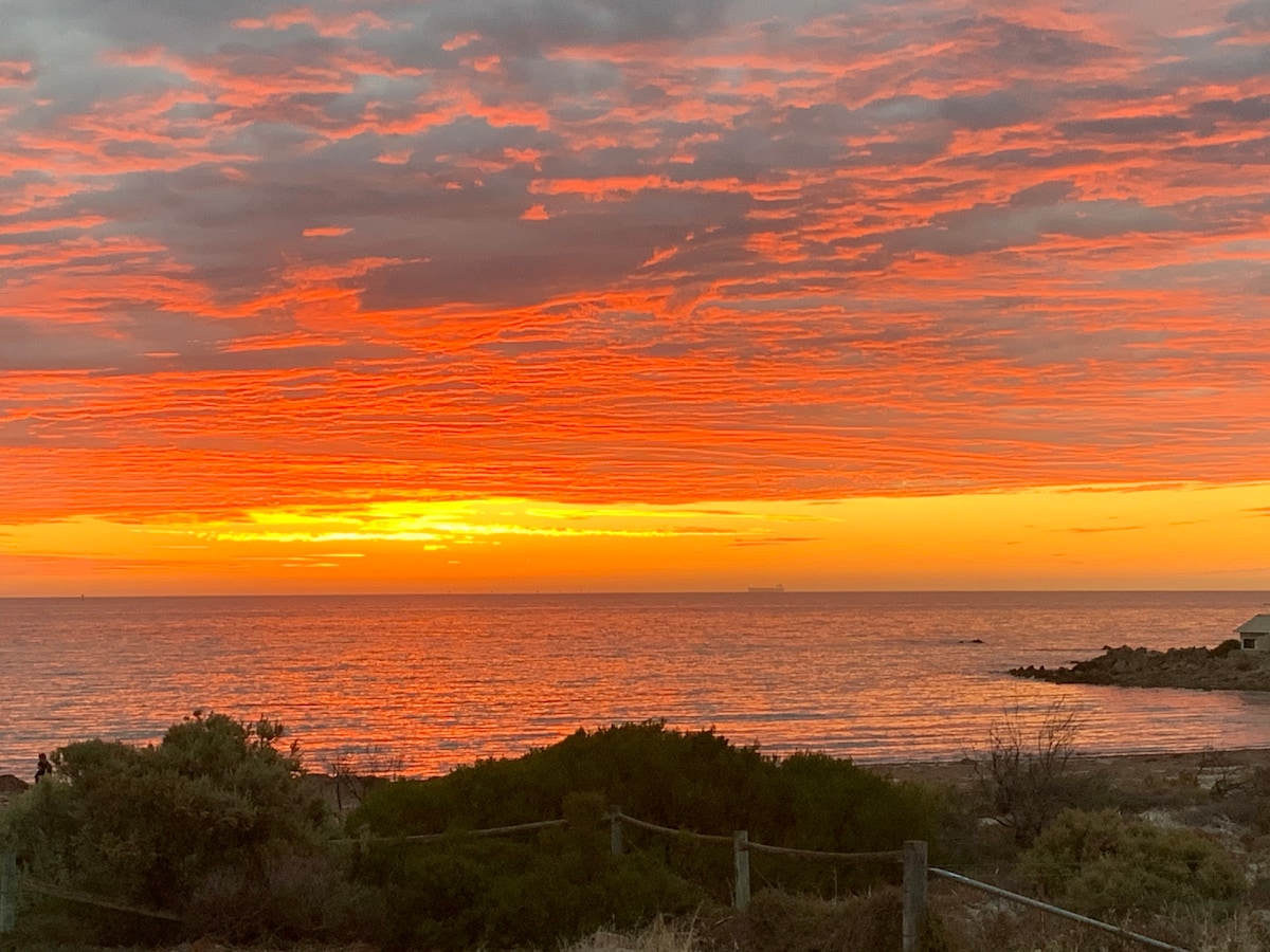 A vibrant sunset paints the sky in shades of orange and red over the calm ocean. The water reflects the warm colors, while silhouettes of bushes and shoreline vegetation are visible in the foreground, creating a serene coastal landscape.