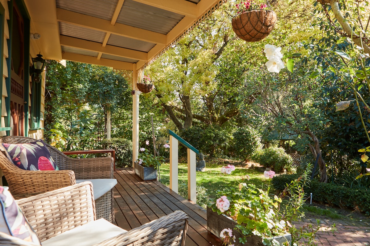 A welcoming porch features wicker seating arranged on a wooden deck, surrounded by lush greenery. Flower baskets hang from the ceiling, and vibrant plants can be seen in pots, creating a serene space for relaxation.
