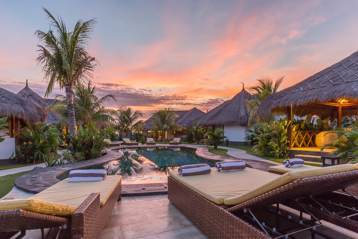 Loungers with soft yellow cushions are arranged around a large pool, reflecting the vivid hues of sunset. Tropical plants and palm trees surround the area, enhancing the tranquil atmosphere created by thatched-roof bungalows in the background.