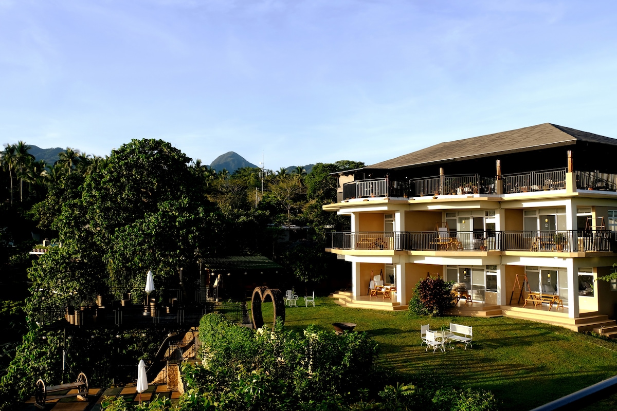The exterior of Tanawin showcases a spacious three-story building surrounded by lush greenery. Balconies with chairs overlook the landscaped garden, featuring several seating areas. A beautiful mountain backdrop completes the scenic view, emphasizing the property’s tranquil setting in nature.
