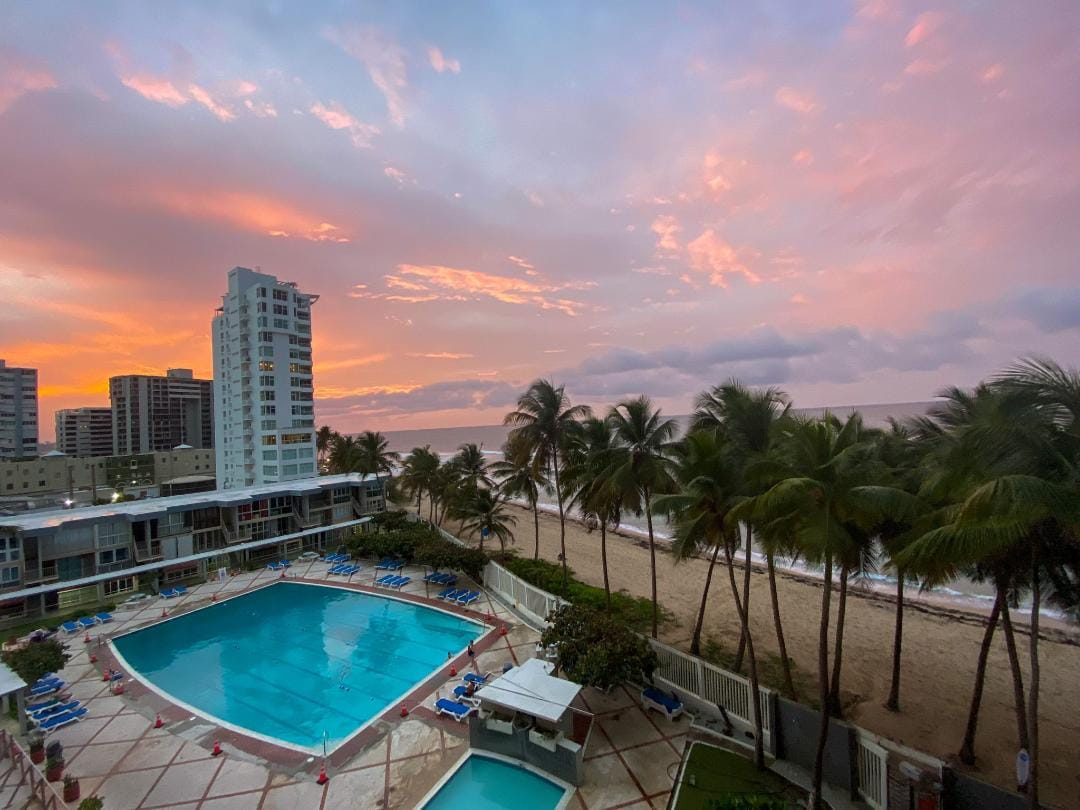 A vibrant sunset creates a colorful sky above a large pool surrounded by palm trees. The beach is visible just beyond the pool, with waves gently lapping at the shore. Nearby buildings are positioned against the backdrop of the ocean.