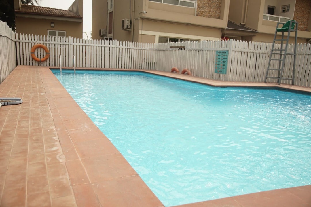 A medium-sized swimming pool is surrounded by a tiled deck. Nearby, a white picket fence provides a boundary, and a ladder leads into the pool. The clear, blue water reflects light, offering a calm and refreshing setting for relaxation.